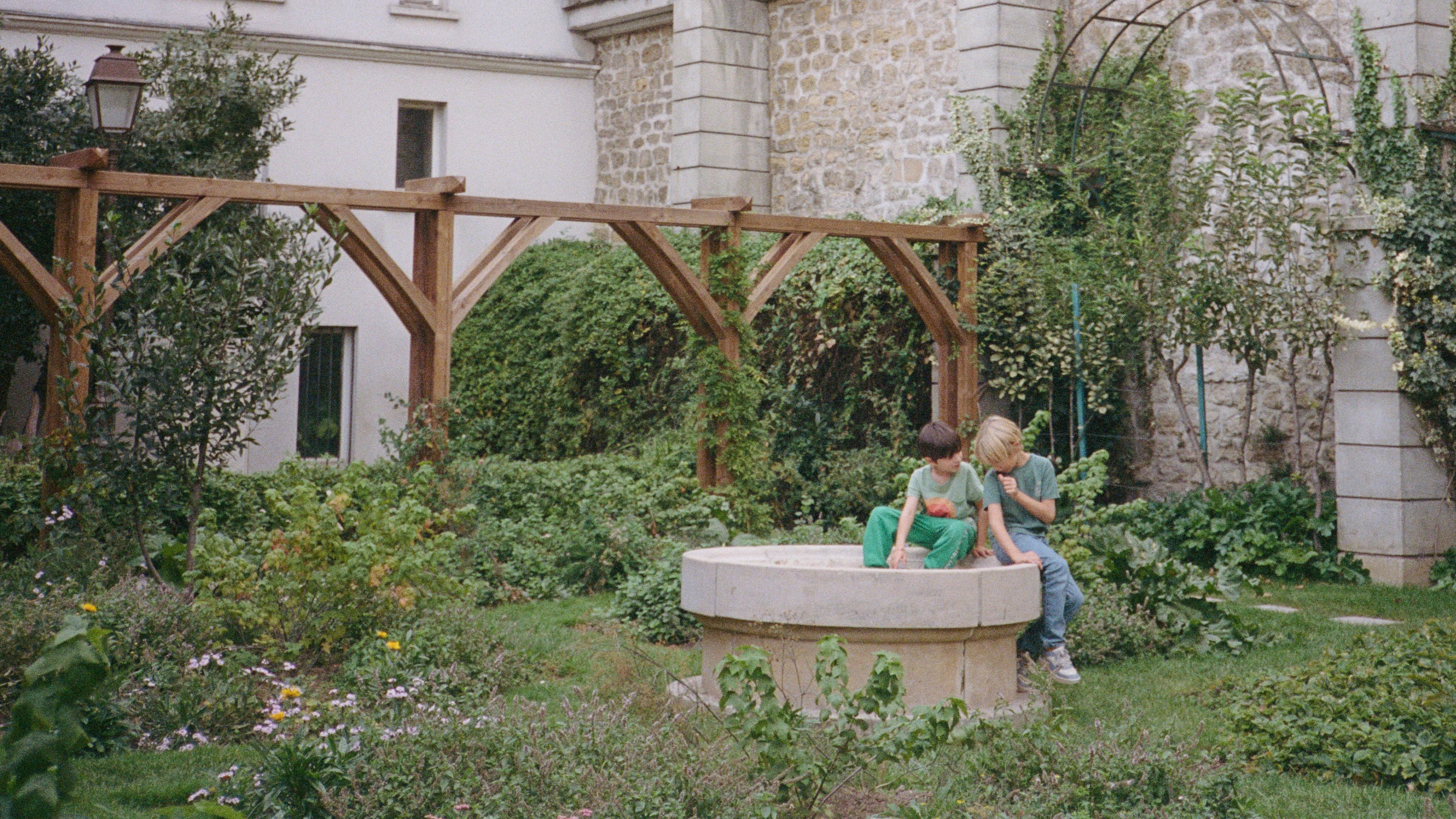 Two children sitting on the edge of a concrete well in a garden with a stone building in the background.